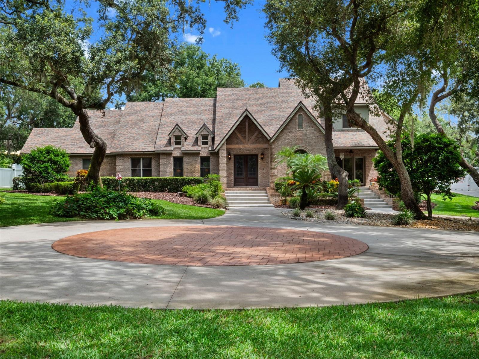 Set on a private cove among towering cypress and moss-draped trees, this extraordinary modern Tudor estate offers a lifestyle of rare beauty and effortless elegance. Nestled in the exclusive Lake Louisa Oaks community—where lakefront living meets timeless design—this custom-built retreat spans over 5,800 square feet of refined living space.
A long, wooded, recently repaved driveway leads to a stately brick façade that opens into a dramatic two-story foyer with honed travertine floors and a spiraling wood staircase that feels like a work of art. Every detail has been thoughtfully crafted—from rich oak trim to natural stone finishes—creating an atmosphere both warm and elevated.
The main level features a serene primary suite, a chef’s kitchen with generous prep and gathering space, a second bedroom suite, spacious laundry room, and inviting living areas—all with sweeping views of the terraced travertine pool deck, complete with rock waterfall, spa, and privately fenced backyard with lush landscaping. Upstairs, three additional bedroom suites, a media room, and spa-inspired baths ensure comfort and privacy for all.
Outdoors, the resort-style design continues with ample entertaining space surrounded by tropical greenery and sculpted stone. Whether hosting under the stars or enjoying a quiet morning by the water, this home was built for both celebration and serenity.
As a resident of Lake Louisa Oaks, you’ll enjoy access to the coveted Clermont Chain of Lakes via private boat ramp and dock on Lake Louisa, along with park-like amenities including a playground, basketball court, and picnic area. The community’s prime location offers the perfect blend of nature, privacy, and proximity—just west of Orlando’s renowned attractions.
Additional highlights include a whole-house generator, private well, and maintained systems that provide peace of mind and year-round comfort.
This is more than a home—it’s a lakeside haven where architecture, nature, and lifestyle come together in perfect harmony.