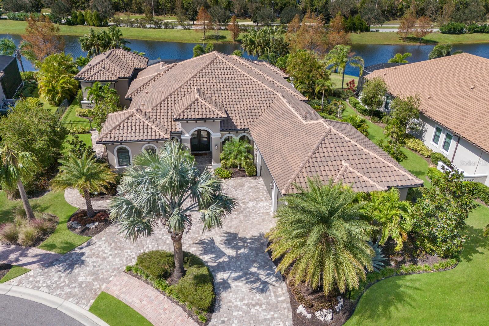 Situated off a cul-de-sac on an incredibly private and serene lake setting, this custom residence in the gated Lakewood Ranch community of Country Club East showcases the addition of a detached CASITA with separate entrance, recently updated interior, a FENCED back yard, IMPACT WINDOWS and DOORS, and even boasts a GENERATOR. Built by John Cannon Homes in 2015, and updated in 2022, this one-story residence encompasses 3,592 square feet of generously open living including a private casita added in 2019, with its own private entrance, full kitchenette, king Murphy bed, living area, full bath, separate washer/dryer, walk-in closet, volume ceilings and stunning lake views.
The expansive outdoor retreat boasts abundant open and under cover spaces, full summer kitchen, PebbleTec heated saltwater pool/spa, panoramic clearview screen, dog run, remote sun screens, speakers, landscape and lanai accent lighting, and a fenced backyard. Ten-foot disappearing impact glass walls seamlessly connect the resort-styled outdoor living space to a large great room with designer feature wall, soaring coffered and tray ceilings, crown molding, porcelain tile flooring and stunning views abound. Chefs will delight in the open kitchen with oversized counter height island, expansive dining area, soft-close dove tail cabinetry, high-performance appliances including Subzero and Wolf, and walk-in pantry.
Relax and unwind in the spacious owner’s quarters offering a spectacular view overlooking the lake, direct lanai access, two separate closets, and a fully remodeled luxuriously appointed bath with double vanity, ample cabinetry, floor-to-ceiling tile, soaking tub, large refreshing shower, towel warmer, and even a morning coffee bar with under counter refrigerator. The main house has two additional guest suites each on separate wings, both with walk-in closets and connected to private full baths. The residence also offers an impressive office behind double-entry doors with built-in shelving and crown molding.
Other notable highlights of this 4,995 total square foot residence includes a conveniently located powder room with direct access from lanai, stunning new designer double entry impact front door, laundry room with cabinetry, sink, and pet door access to the dog run, two tankless water heaters, 2024 and 2019 AC units, instant hot in the kitchen and casita, impact windows and doors, elevated landscaping with lighting, circular pavered driveway, and an oversized three car garage with overhead storage. Country Club East provides its residents with the privacy of gated-community living, three community pools, and fitness center, all within a renowned community infamous for its vibrant Lakewood Ranch lifestyle near dining, shopping, polo, golf, tennis, community events and still close to the UTC area, the beaches of Sarasota, and, of course, in an A-rated school district near Out-of-Door Academy and just a few miles from IMG. Residents also have the non-mandatory option to become members of the Lakewood Ranch Golf and Country Club, which offers four championship golf courses, two clubhouses, tennis, pickelball, fitness center, heated pools, full social calendar and so much more.