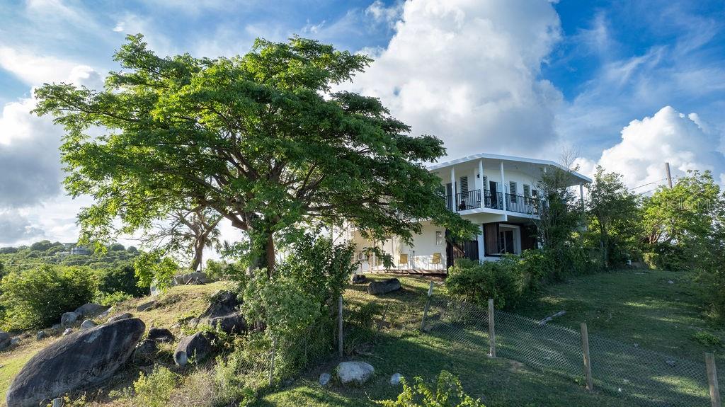 For the first time on the market, this unique two-unit home sits on 3 private acres in the highly sought-after La Llave neighborhood. Accessed by a winding uphill driveway, the property is positioned at the highest point of the hill to offer unobstructed 180-degree views of the southeast of Vieques, Sunbay and the keys of La Esperanza. The serene setting is a true escape from the everyday, blending tranquil nature with stunning vistas.
The home is divided into two independent units, each with its own private entrance, making it perfect for multi-family living, vacation rentals, or as a versatile investment property. Both units feature spacious, open-concept floorplans flooded with natural light and designed to maximize the scenic views.
The upper unit welcomes you with a large wraparound balcony that offers sweeping views of the Caribbean Sea. Inside, a vast great room combines living, dining, and a fully-equipped kitchen, all designed for entertaining and relaxed living. This unit includes three bedrooms, with the principal bedroom offering an en-suite bathroom, as well as an additional full guest bathroom.
The lower unit features an expansive open living, dining and kitchen area, three comfortable bedrooms, and one full bathroom. The layout provides flexibility, allowing for easy division of the space to create a carport or additional storage area.
The property is surrounded by mature trees that provide natural shade and a refreshing breeze, creating the perfect spot to relax in a hammock and enjoy the tranquil surroundings. The vast 3-acre lot offers ample room to build a pool and terrace, as well as create a stunning garden. To the rear of the property, a beautiful mountainous landscape dotted with fruit trees provides plenty of space for further expansion, additional structures, or even agricultural pursuits.
This home offers tremendous potential, whether you seek a private home, an investment property, or a combination of both. With its breathtaking views, spacious accommodations, and prime location (in proximity to both urban centers of Vieques), it is poised to become one of the most desirable properties on the island.