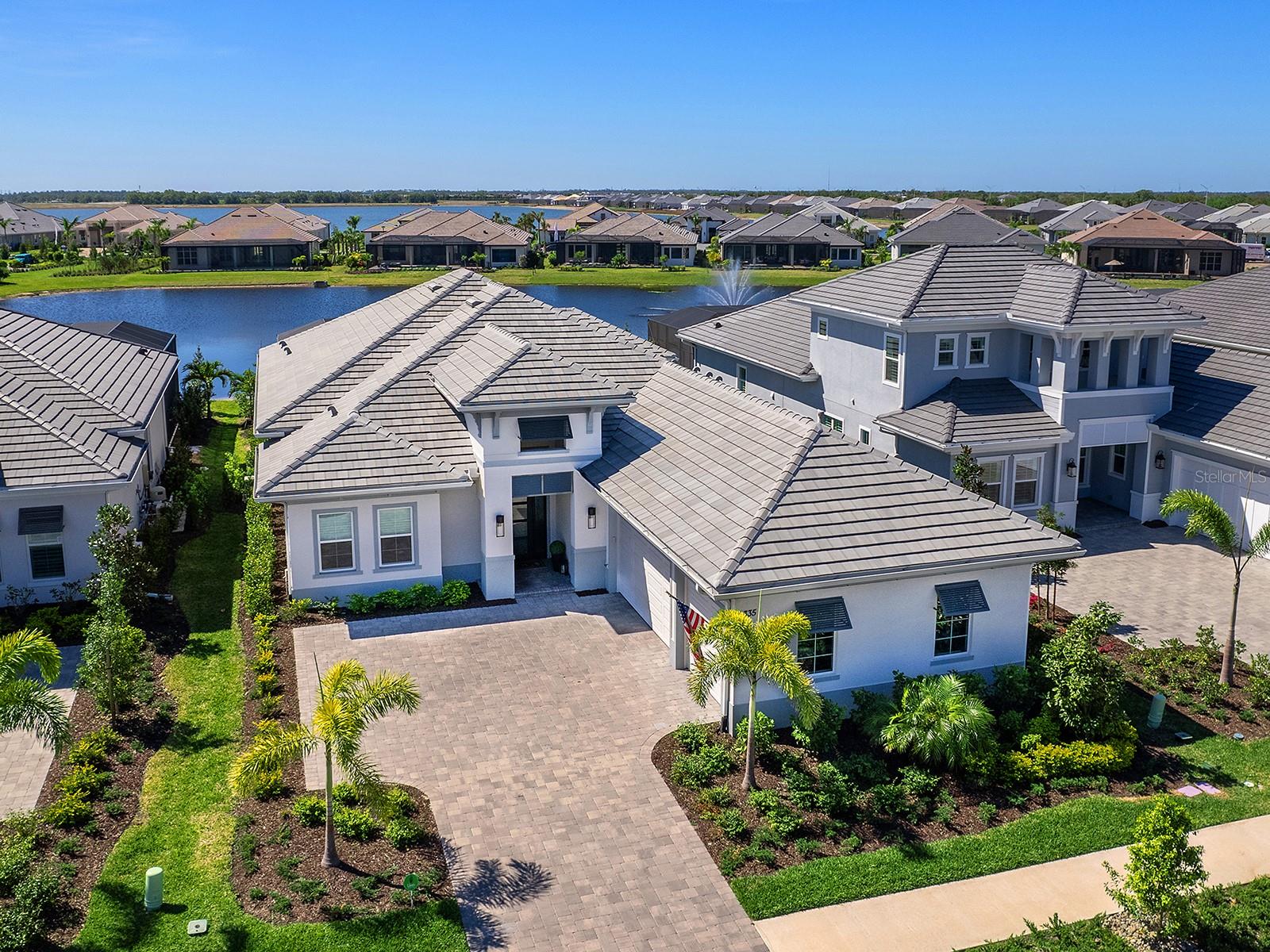 Offering an exquisite blend of coastal elegance and modern functionality, this maintenance-free home is set against the serene lake with a stunning fountain view. An oversized paver driveway leads to the home’s inviting coastal elevation, enhanced by professionally designed landscaping, coach lighting and decorative shutters that accentuate the entrance. Impact glass throughout ensures peace of mind, while wide plank wood-look tile floors bring style to the main living spaces. Stepping through the custom Aquatek® glass front door with an iron frame, the foyer welcomes with a tray ceiling and designer chandelier. The open concept seamlessly blends the main living areas, creating an ideal layout for everyday living. An illuminated floating tray ceiling with custom trim adds ambiance to the living room while the wall of pocketing sliders leads to the lanai. The dry bar, situated between the living and dining area, features glass upper cabinets, quartz countertops, a beverage cooler, and a tile backsplash. Custom-trimmed walls define the dining area, with plenty of space for entertaining. The kitchen is a dream, boasting quartz countertops, a spacious floating island with seating and storage, stainless steel KitchenAid appliances including a five-burner gas cooktop, and a glass-front corner cabinet. Soft-close cabinetry, a pantry cabinet with pullout shelving and a large window with remote blinds complete the space. The primary suite is a retreat with a tray ceiling with a designer chandelier, two custom walk-in closets and French doors opening to the lanai. The en-suite bath is an oasis, featuring a free-standing soaking tub, an expansive walk-in shower with designer tile flooring, a private water closet and a double-sink vanity with quartz countertops. Three additional guest bedrooms provide ample accommodations, with one offering an en-suite bath and the others sharing a well-appointed bath with a walk-in shower and custom vanity. French doors open to a refined office space with a picture window, tray ceiling and beautiful chandelier. Designed for relaxation, the outdoor space is unparalleled. The enormous paver lanai is plumbed for an outdoor kitchen and fireplace, with a covered living and dining area under a tray ceiling with recessed lighting. An outdoor kitchen will be installed in July of 2025, ready for alfresco dining with a view. The resort-style pool includes sunken spa, fountain bowls, a panoramic screen for uninterrupted lake views and a sun shelf for lounging. A dedicated pool bath adds convenience. Additional highlights include a recirculating hot water system, laundry room with white cabinetry and subway tile backsplash, a three-car garage with epoxy flooring, and a powder room with a pedestal sink and lighted mirror. Wild Blue offers an unparalleled lifestyle with a resort-style clubhouse featuring concierge service, a theater, fitness studio, golf simulator, billiards and an expansive pool complex with two resort-style pools, a restaurant and a patio bar coming soon. Outdoor enthusiasts enjoy scenic walking trails, a yoga lawn and a putting green, while sports lovers have access to the Midway Sports Complex with eight pickleball courts, six tennis courts and a professional-grade pro shop. A dog park and playground further enhance the community’s appeal. Ideally near The Mall at UTC, Lakewood Ranch Main Street, Waterside Place and top dining destinations, this home embodies the pinnacle of luxury living in Lakewood Ranch.