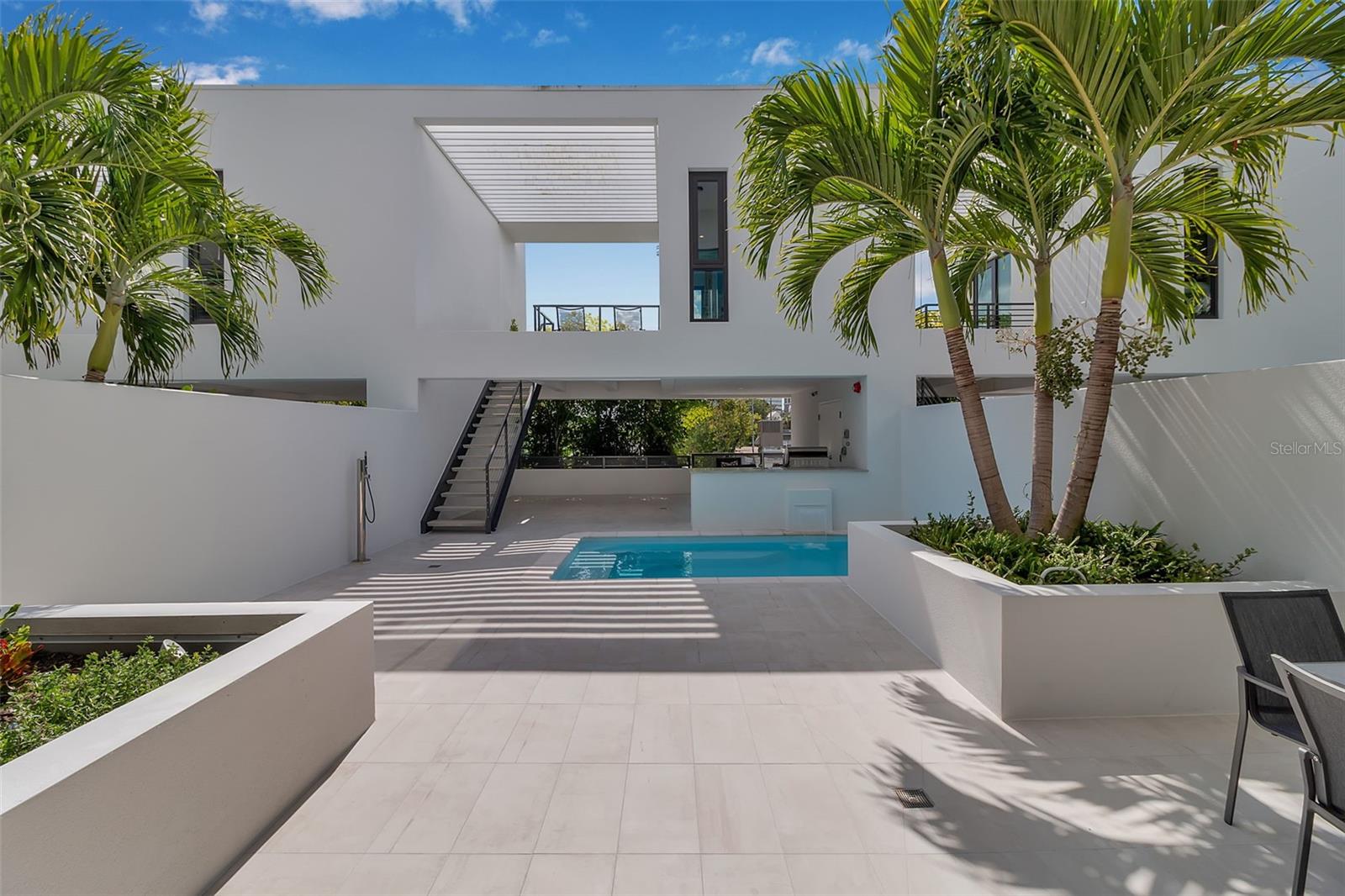 Anchored in Sarasota’s vibrant Rosemary District, a hub of art, dining, and design just steps from downtown, this contemporary three-story residence captures the essence of urban coastal living. Defined by its clean architectural lines, striking white façade, and geometric precision, it offers a rare balance of privacy and proximity in one of Sarasota’s most walkable neighborhoods. A sleek iron-and-glass entry framed by textured tile sets the tone for what’s inside: light, volume, and effortless connection to the outdoors. The floating staircase, with open risers and maple treads, becomes a sculptural centerpiece beneath ceilings that soar up to 13 feet. A three-stop elevator ensures convenience from the ground-level garage to the uppermost terrace. The main living level unfolds with gallery-like openness, a seamless blend of style and function. The sleek kitchen, featuring quartz countertops, premium stainless-steel appliances, and clean-lined cabinetry, overlooks the dining and great room. The open living space is complete with expansive sliders open directly to the courtyard and its tranquil Zen pool, where the rhythm of water and greenery softens the modern form. With its summer kitchen, fire table, and covered dining terrace, the home transitions effortlessly from quiet mornings to lively evenings. Upstairs, the primary suite feels like a retreat, complete with a glass-framed balcony, spa-inspired bath, and thoughtful details that invite calm. Two additional guest rooms, one that is currently used as an office, mirror the home’s minimalist aesthetic and abundant natural light. A private guest casita, accessible from the courtyard, expands the home’s flexibility. With a kitchenette, full bath, and its own balcony, it’s ideal for visitors, another primary suite or a fitness space, all with the independence of a separate entry. Constructed of concrete and steel with impact-rated glass, every element underscores quality and low-maintenance design, perfect for those seeking the ease of a lock-and-leave lifestyle. Positioned in Sarasota’s coveted Rosemary District, one of Sarasota’s most walkable neighborhoods, this residence places the best of the city just outside your door. Stroll to downtown dining, art galleries, live music, and cultural venues, or stop by the weekly Sarasota Farmers Market. Boutique shops, theaters, and cafés line the nearby streets, while the beaches, the John Ringling Bridge, St. Armand's Circle, and Sarasota Bay are only minutes away. With convenient access to Sarasota Bradenton International Airport, this address perfectly blends urban luxury, outdoor living, and strong architectural design, offering a downtown modernism that feels both refined and deeply livable.