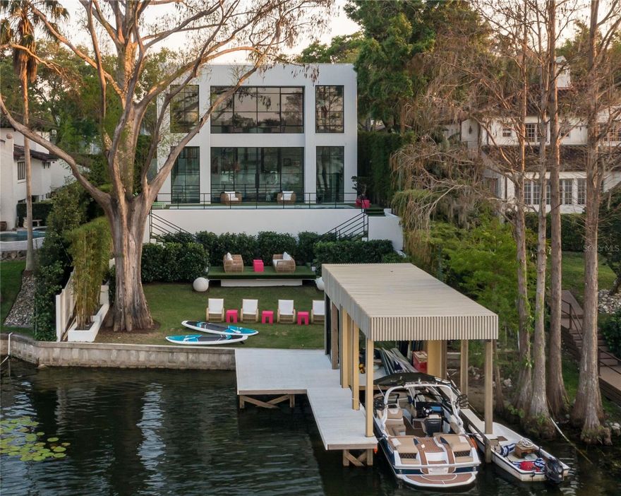 An architectural triumph, this one-of-a-kind modern masterpiece was custom designed and built in 2023 by the award-winning Z Properties and is ideally sited on the shores of Lake Osceola, part of the Winter Park Chain of Lakes. Radiating bold sophistication and contemporary elegance, the residence is a curated work of art, where floor-to-ceiling glass walls and striking steel windows and doors flood the interiors with natural light and deliver unobstructed panoramic lake views from nearly every room. Enter through a dramatic 12-foot glass pivot door into a 21-foot-long art gallery foyer adorned with a sculptural linear chandelier and Venetian plaster walls and ceilings that lend a soft, refined texture. The open-concept living spaces are accentuated by terrazzo floors, 12-foot ceilings, and thoughtful spatial design—including a great room with a sitting area and dining space, a separate media room concealed behind custom 12-foot decorative doors, and a sleek galley-style kitchen outfitted with fully integrated Miele appliances and a secondary glass pivot door for seamless indoor-outdoor flow. Ascending one of two custom staircases, the second floor features warm hardwood floors and a showstopping primary suite that feels suspended above the lake, backyard and pool. This serene retreat offers an all-glass shower, spa-inspired soaking tub, beautifully finished bath cabinetry, and an upholstered wall that subtly conceals a walk-in closet and built-in storage. A second bedroom suite offers an inspired use of space with clever screen walls for privacy and flexibility. Detached from the main home, a third bedroom suite doubles as a guest house, home office, or gym, complete with an en-suite bath, custom built-ins and a wet bar. Outside, the terraced backyard is a tranquil oasis with a saltwater pool and spa, turf lawn for year-round perfection, dual staircases leading to a sundeck and a private, covered boat dock crowned by a glass-enclosed studio mirroring the home’s signature design. Bamboo privacy walls, soaring palms, and a Banyan tree imported from South Florida create a lush and artful landscape. The two-car garage includes epoxy flooring and dual EV charging stations. A singular residence in every sense, this home blends innovative architecture, luxurious finishes and thoughtful design into a rare offering unlike anything else in Winter Park. Built by Z Properties, celebrated in Florida Design and numerous design publications, this is a home for those who appreciate beauty, intention and the art of living well.