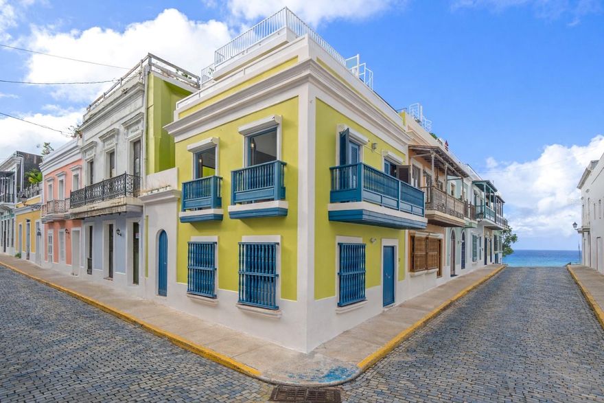 Situated on one of the most iconic and photographed blocks in Old San Juan, at the corner of San Sebastián and San Justo Streets, this distinctive corner townhouse forms part of a visually unified row of privately owned homes.  Distinguished by its corner placement and elevated setting, the residence anchors the block with a vivid canary-green façade accented by cobalt-blue doors, windows, and wood balconies—an architectural palette emblematic of Old San Juan’s historic character.  Its position on one of the highest points of the old city affords constant trade-wind cross breezes and open outlooks, with ocean and bay glimpses from multiple levels.  

The home occupies an intimate footprint, yet unfolds across three levels designed to maximize light, airflow, and indoor-outdoor living.  The ground floor features an open kitchen, dining, and living area ideal for entertaining, opening onto a private interior courtyard.  A signature outdoor shower—framed in patterned Spanish-style tiles and open to the sky—serves as both a functional amenity and a classic Old San Juan architectural detail, accessed discreetly from within the home and directly from San Sebastian street through an arched blue door.  

The second level offers two bedrooms and a full bathroom.  One bedroom opens onto a wide balcony facing San Justo Street, capturing ocean glimpses and prevailing breezes, while both bedrooms feature Juliet balconies facing San Sebastián Street, enhancing natural light and cross ventilation throughout.  The third level hosts a spacious flex room with a full bathroom—suitable as a primary bedroom, family room, or studio—opening directly onto an L-shaped terrace with expansive views over the historic rooftops, the bay, and the Atlantic Ocean beyond.  

A spiral staircase ascends from the terrace to an open rooftop deck outfitted as a relaxed lookout and lounge area.  From this elevated vantage point, the home commands sweeping panoramic views of Old San Juan’s skyline, coastline, and surrounding cityscape.  Located on San Sebastián Street, one of the district’s most celebrated thoroughfares and within close proximity to renowned restaurants, cafés, and cultural landmarks, this exceptional corner townhouse offers a rare opportunity to own a visually iconic, light-filled residence that captures the essence of Old San Juan’s architectural and cultural legacy.