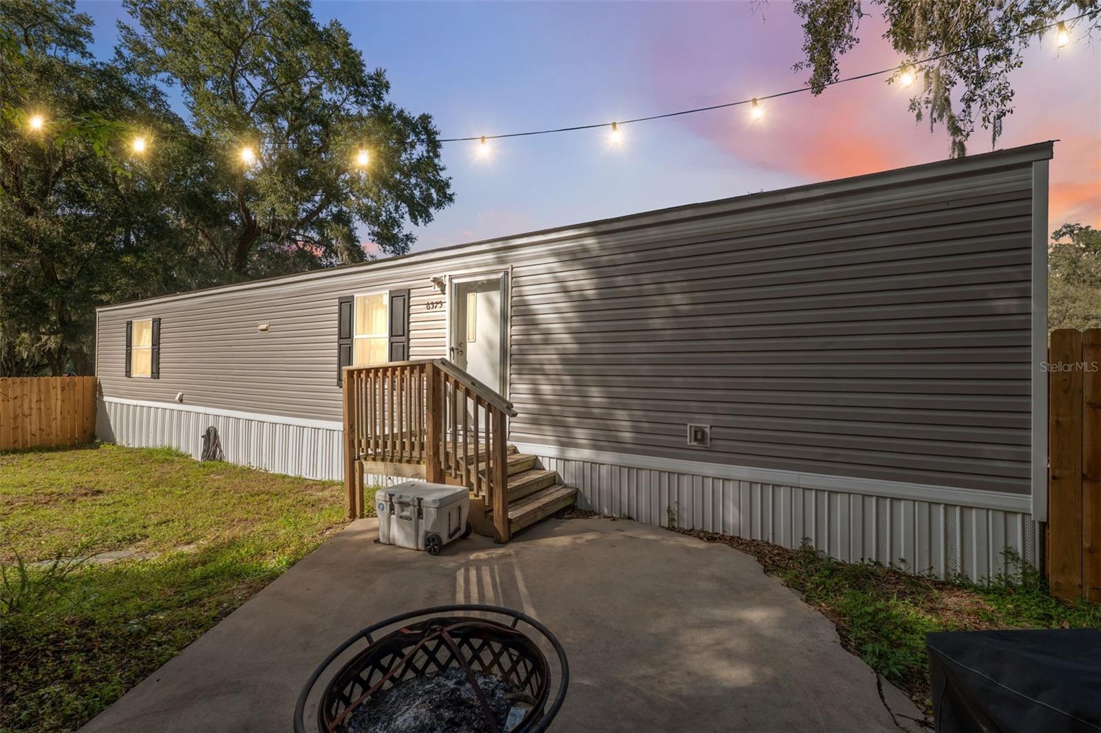 Set amid a peaceful, lightly wooded neighborhood, this charming mobile home offers a comfortable and inviting retreat surrounded by nature. With two bedrooms and two full bathrooms, the home blends cozy living with practical design in a serene setting. The exterior features light gray vinyl siding with white skirting, a paved drive with a gravel extension for additional parking, and a spacious front yard shaded by mature trees that add privacy and a natural touch. Step inside to an open-concept living room filled with soft natural light from multiple windows, complemented by recessed lighting, a ceiling fan, and neutral finishes that make the space feel airy and welcoming. The adjoining kitchen showcases a functional U-shaped layout with wood-finish cabinetry, light laminate countertops, and a seamless connection to the living area—perfect for everyday convenience. The dining area features warm-toned laminate wood flooring, recessed lighting, and a glass-paneled door that leads outside, allowing easy access for morning coffee or evening relaxation. The primary bedroom offers a peaceful retreat with laminate flooring, beige wall panels with dark trim, and direct access to a private en suite bathroom featuring wood-pattern vinyl plank flooring and soft overhead lighting. The second bedroom and full bathroom are equally comfortable, providing versatility for guests or a home office. A separate laundry room adds extra functionality with wood-pattern flooring and ample space for storage. Outside, the rear entry features a small wooden staircase and railing leading to an open backyard surrounded by greenery, offering plenty of room for outdoor activities, a garden, or a quiet sitting area to enjoy the Florida breeze. The home’s lightly wooded surroundings create a sense of privacy while still being part of a friendly residential area. This property is conveniently located close to The Villages, where residents can enjoy shopping, dining, and year-round entertainment. Also nearby is Carney Island Recreation and Conservation Area, featuring nature trails, picnic areas, and fishing spots along scenic Lake Weir, and Lake Weir itself, known for its clear waters, boating, and lakeside restaurants. Offering both peaceful country living and access to nearby conveniences, this mobile home provides the perfect balance of comfort, simplicity, and connection to the natural beauty of Central Florida.