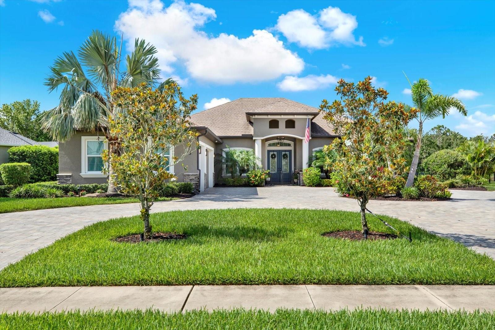Welcome to this exquisitely upgraded “Former McKenzie Oasis Model” home, perfectly nestled in the serene community of Twin Rivers. Offering 3 bedrooms, 3 bathrooms, a den, and a flexible bonus/media room, this home seamlessly combines refined living with thoughtful updates throughout. A spacious 3-car, climate-controlled garage with upgraded storage and LED lighting ensures year-round comfort and functionality.
Set back on an impressive paver driveway, this home boasts an extended U-shaped layout for ample parking and elegant curb appeal, enhanced by $25,000 in lush landscaping, mature trees, and a custom outdoor water feature that creates a welcoming ambiance. The front porch is gated for added security and charm, while the paver walkway leads to a beautifully landscaped side yard. Inside, you’ll find new laminated wood flooring in all bedrooms, crown molding and can lighting throughout, plantation shutters, and a stunning stonewall fireplace. Soaring 11’4” coffered and tray ceilings and new ceiling fans in every room elevate the sense of comfort and style. The gourmet kitchen is a chef’s dream, boasting new backsplash, solid wood cabinetry, high-end granite and cultured marble countertops, stainless steel appliances, a pot filler, reverse osmosis water treatment at the sink, a spacious 10’ island with new light fixtures, and two walk-in pantries. Glass office doors, upgraded Kohler toilets, bidet toilet seats, and a complete remodel of the pool bathroom further enhance the home’s refined details. The owner’s suite is a serene retreat with direct access to the lanai, dual custom closets, and a luxurious en suite bath with double vanities, a soaking tub, and a walk-in shower. The bonus/media room offers direct lanai access and flexible use as a guest suite or additional living area, perfect for multigenerational living. Step outside to your private oasis: an 18,000-gallon saltwater pool with upgraded LED lighting, a lap lane, waterfall feature, and electric heater—all remotely controlled. Pool lanai blinds and upgraded lanai lighting create a resort-style atmosphere, ideal for year-round enjoyment. An outdoor kitchen with grill and refrigerator adds to the home’s impressive entertaining options. Additional feature include: New pool pump, Attic lift with remote control (dumb waiter), New air conditioning in the garage, House pre-wired for generator, Complete ADT security system with hardwired cameras throughout the interior and exterior perimeter, upgraded landscape lighting with timer, Sprinkler system adjusted and expanded with a new WiFi-controlled panel for easy management, New outdoor light fixtures, New front load washer and dryer on stands. Twin Rivers offers an unparalleled lifestyle with parks, nature trails, playgrounds, and kayak launches—all with low HOA fees and no CDD. Located in a no-flood zone (X500) and move-in ready, this home perfectly blends luxury, efficiency, and multigenerational living in a sought-after community. Don’t miss this rare opportunity to own a beautifully upgraded, move-in ready home. Contact us today to schedule your private showing!