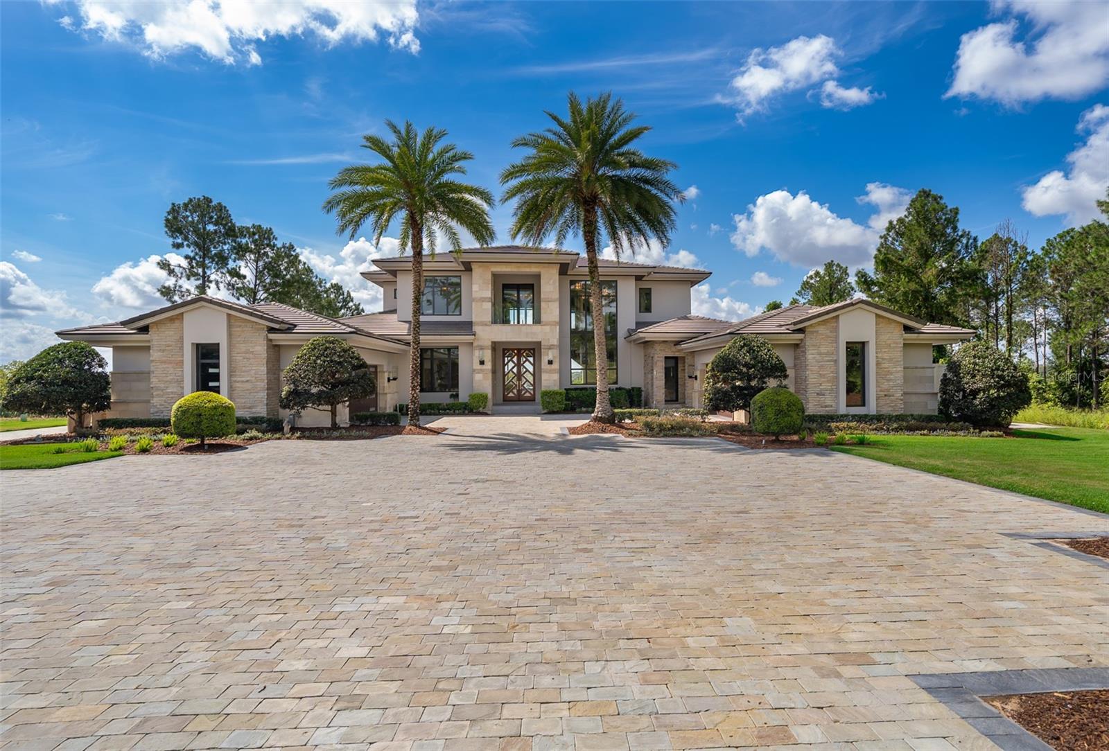 5 Bedrooms | 5 Full Baths | 3 Half Baths | Lake front-Private Pool | 4-Car Climate-Controlled Garage
Experience refined living in this custom-built, lakefront ( Lake Apopka) estate in the exclusive, guard-gated community of Bella Collina. Designed for both grand entertaining and everyday luxury, the open-concept main living area features soaring ceilings, a stone fireplace wall, and expansive sliding glass doors that blur the line between indoor and outdoor living. The chef’s kitchen is a true showpiece with a waterfall-edge quartz island, Thermador appliances, custom cabinetry, and a spacious walk-in pantry—perfect for hosting gatherings or preparing intimate meals.
The main level boasts a custom-built bar with dual wine refrigerators, an entertainment lounge that flows seamlessly to the lanai, a home office, a guest suite with en-suite bath, and two designer guest baths. The luxurious primary suite is a private retreat, featuring a stone fireplace, morning bar with Thermador coffee system, spa-style bath, and private lanai with fireplace—ideal for unwinding while overlooking the serene pool and landscaped grounds.
Upstairs offers three additional en-suite bedrooms boasting balcony access, perfect for enjoying morning coffee or evening sunsets, a bonus/media room, and half bath, ensuring comfort and privacy for family and guests alike. Outdoors, the home transforms into a resort-inspired retreat with a fully equipped summer kitchen, covered lounge and dining areas, and multiple spaces designed for both relaxation and entertainment. Host sunset gatherings poolside, enjoy weekend brunch under the wood-paneled lanai, play golf in your private putting green with bunkers from an elevated 70-yard (approx.) tee or take in the views while lounging by the sparkling pool framed by lush landscaping.
Premium upgrades include a Crestron Home Automation System which allows the control of lights, shades, temperature, music, security and more from a single platform, elevator, built-in speaker system, water filtration, and a climate-controlled 4-car garage with utility area. Located within a short distance to the amenity-rich setting of Bella Collina, featuring a Nick Faldo Championship golf course, a luxurious spa, Har-Tru tennis courts, pickleball courts, clubhouse, outdoor pool complex, cutting-edge fitness center with complimentary fitness classes, lake views and monthly culinary and/or wine events. Don’t miss the opportunity to own this one-of-a-kind luxury retreat in Central Florida’s premier private community.