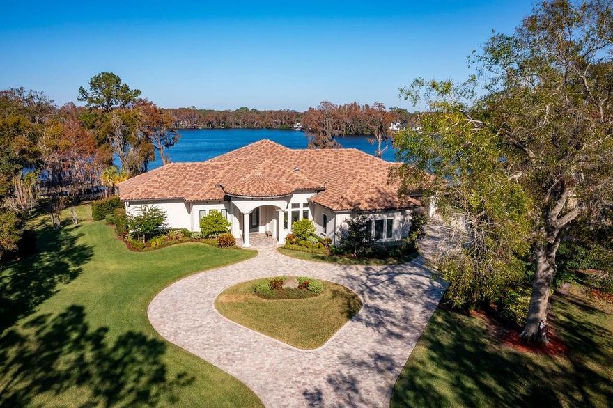 A harmonious blend of contemporary elegance and lakefront serenity, 9903 Menander Wood Court presents an exceptional opportunity to live the enviable boating lifestyle. With 143 linear feet on Pretty Lake, a highly coveted ski lake in the Tampa Bay area, the home can accommodate a boat dock and ramp, welcoming endless recreational pursuits. Upon arrival, the circular driveway exudes a sense of stately grandeur, weaving through a picturesque one-acre lot with extensive landscaping. The impressive iron double doors provide an elegant entry revealing a spacious, light-filled vestibule that’s adorned with spectacular one-of-a-kind custom light fixtures and beautiful lake views. The well-designed floor plan features 12- to 14-foot ceilings, solid 8-foot mahogany doors, and porcelain tile flooring throughout the main living areas, all in a mesmerizing one-story design of over 7,500 square feet.  Designed with entertaining in mind, the kitchen is outfitted with an impressive 13-foot center island, custom wood cabinetry with pull-outs, quartz countertops, an expansive pantry with appliance bar and state-of-the-art appliances that include double ovens and a six-burner induction cooktop. This culinary haven flows effortlessly into the great room, where 12-foot sliding glass doors frame panoramic views over the lake. A tranquil respite, the primary suite features double walk-in 14-by-9-foot closets, and a spa-like bath with dual vanities, a soaking tub overlooking the lake, custom-designed mirrors and a large shower with rain head plumbing. Two separate guest wings include 3 more opulent bedrooms, all of which feature en-suite baths and captivating views of the glistening lake. The spacious home office is adorned with floating mahogany shelves and automatic blinds, while the theater room is equipped with a 10-foot media screen, surround sound and electric seating. Outside, restful retreats or large gatherings, await on the out-sized 900-square-foot terrace, featuring an elegant tongue-and-groove wood ceiling, paver decking, automatic retractable screens and a pool bath. In addition, this exterior space has been thoughtfully pre-plumbed for an outdoor kitchen and pizza oven. The 1+ acre homesite provides copious space for a newly designed resort-style pool set against the breathtaking backdrop of the lake. The lovely white sand beach provides plenty of space for water activities or just relaxing with family and friends at the water’s edge. Further exterior amenities include a kayak rack, a fenced dog run and room for a boat ramp. The property includes an extensive solar power system, Control4 home automation system, energy-efficient Icynene insulation, storm-rated windows and doors, two spacious two-car garages with Tesla power walls, and a water filtration system. Offering unparalleled luxury living in an unrivaled lakefront setting, this Northbridge at Lake Pretty home is available for private showings by appointment.