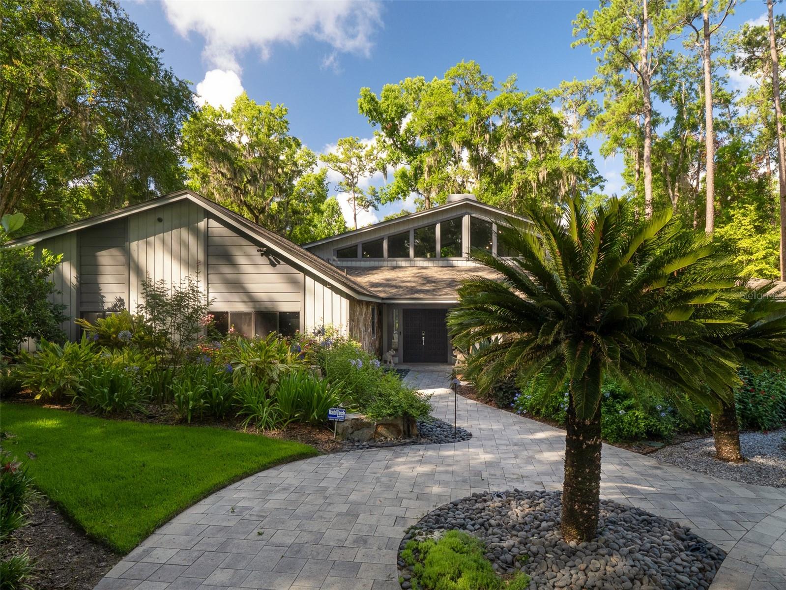 Architectural Sophistication on 1.1Private In-Town Acres~ Privately situated in the desirable Hermitage neighborhood, this architecturally distinctive residence on 1.1 beautifully landscaped acres offers a rare blend of scale, serenity, and convenience. Recently updated, this sophisticated property balances design-forward style with everyday elegance. Enter through double doors to a sunlit interior featuring travertine floors, soaring ceilings, and floor-to-ceiling windows that seamlessly connects indoor and outdoor living. The expansive great room, anchored by a dramatic fireplace, flows effortlessly between multiple entertaining and living zones, including a custom bar area with backlit quartzite—all overlooking the newly-built (2025) resort-style pool and private backyard. The great room opens to the chef’s kitchen, as functional as it is beautiful, with dual prep stations, a waterfall edge countertop, induction cooktop, steam and convection ovens, and sleek custom cabinetry. The serene primary suite offers a spa-like retreat with a soaking tub, large walk-in shower, and custom closet. The private secondary wing includes two bedrooms, a shared bath, a flex/media room, and a separate gym. ~
Recent enhancements include new flooring, impact-rated windows and updated lighting that elevates both ambiance and energy efficiency. Outdoors, a screened-in pool area is enhanced with water features and surrounded by a grill room with a summer kitchen, a full pool bath, multiple patios, fountains, a remarkable fire table, and a putting green. The park-like backyard private oasis is nothing short of magical - an illuminated garden path winds past an additional detached 1BR/1BA studio cottage with kitchenette, weaving through extensive landscaping, fruit trees, and a vibrant tapestry of flowering shrubs and greenery. ~ The exterior lighting is thoughtfully designed for versatility—offering soft, ambient illumination for everyday elegance, while effortlessly transforming into a vibrant, colorful display to elevate special occasions, festive gatherings, or celebratory events. ~ Equipped with smart home integration and a state-of-the-art security system, this home offers intuitive control of lighting, climate, sound, and surveillance. Additional highlights include a three-car garage, spacious laundry room, and concrete block construction—all just minutes from The University of Florida, top medical centers, and Gainesville’s finest dining and shopping. https://gainesville360.com/2025/2261/