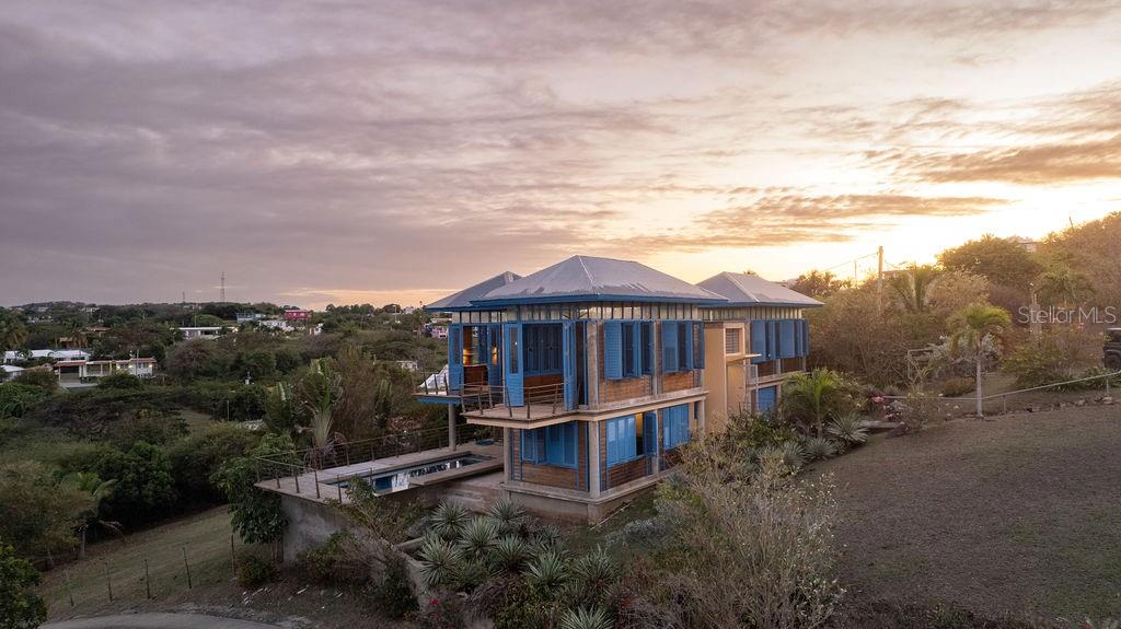 This exclusive house inspired by traditional turn-of-the-century Puerto Rican architecture, was designed by renowned architect Pablo Ojeda, whose work has earned international recognition, including the 2001 URBE Prize for Architectural Excellence in Design and Construction.
Ideally situated atop a hill at the end of a private road, this home offers constant breezes throughout and sweeping sea views over the lush tropical foliage of the valley below. The main island, Culebra and St. Thomas can be seen dotting the horizon from multiple parts of the house. Included in the sale are two contiguos lots which, together with the rest of the land, total 1.25 acres, adding an element of seclusion to the location. Such acreage is a rare find in the neighborhood of Bravos de Boston which is coveted for, among other things, its proximity to the sea, the ferry, and the shops, restaurants and cafes of Isabel II.
The house’s design embraces traditional materials and construction methods reimagined for modern tropical living. Intentionally oriented to take advantage of the ocean views and sea breezes, t’s massive blue French doors and louvered windows open up to various outdoor balconies and living spaces, blurring the line between indoors and outdoors. The surrounding mature gardens and plantings in the central courtyard complement the architecture and bring nature into the heart of the home. Polished cement floors and countertops add a contemporary touch while being easy to maintain. The soothing sound of the fountain coupled with the backdrop of the ocean waves and local birds, envelops you in the serene tropical landscape as you relax by the pool.
The house can be accessed by a quaint pedestrian pathway via the courtyard or via the parking area over an elevated walkway that leads to a split-level entryway. Spanning approximately 2,000 sqft, the home is spread across two levels, with the main living areas on the upper level and guest and communal spaces on the lower level.
On the upper level, the dining area, which has both sea and courtyard views, leads into the kitchen, which has ample counter and storage space and room for an island or small breakfast nook. Off the kitchen is a sea-facing balcony; a perfect place for a morning coffee or evening cocktails. The adjacent living room features soaring ceilings and clerestory windows that allow natural light to seep in. At the end of the living room in another sea-facing balcony. To the rear is the master suite with an ensuite bathroom, including a luxurious soaking tub. The upper level also includes a half-bathroom which can also accommodate a washer/dryer.
The main focal point of the lower level is the flora-filled courtyard terrace. Toward the back-end of the property is a large bedroom with an ensuite bathroom, which can also be accessed directly from the adjacent terrace. This bedroom has an outdoor seating area accessible through a separate entrance. Toward the front is an ocean-facing the tiered pool area, which has ample room for a shaded dining area. Directly adjacent is a guest suite and kitchenette, with accordion doors that open up to the pool. A storage room with plumbing provides the potential for easy conversion into a third full bathroom.
The grounds include a spacious parking area bordered by olive trees, a variety of palms, oleander, cactuses, and tropical plants. The house is just a two-minute walk to the pristine waters of La Chata beach and a four-minute drive to town.