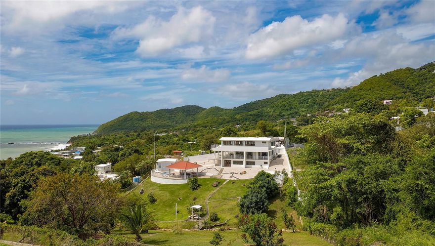Set on an expansive 1.1-acre homesite within El Cocal Country and Beach Club, this private hillside residence captures sweeping ocean views and a sense of quiet retreat that is increasingly rare along Puerto Rico’s coast. Thoughtfully positioned to embrace both the surrounding greenery and the Caribbean beyond, the home offers a seamless balance between indoor comfort and outdoor living. The main residence unfolds with an airy, open-concept design where the living room, dining area, and kitchen connect effortlessly to a generous covered terrace. Framed by wide openings and clean architectural lines, these spaces are defined by natural light, cooling breezes, and uninterrupted views of the sea to create an ideal setting for both relaxed daily living and elegant entertaining. The bedroom layout is both functional and refined. Two guest bedrooms and two full baths, one ensuite, are located on the main level, offering comfort and privacy. Above, the primary suite occupies its own level, designed as a true retreat with a walk-in closet and a private terrace that opens to panoramic ocean vistas, an intimate space for quiet mornings or sunset evenings. A separate lower-level apartment adds versatility to the property, complete with its own bedroom, ensuite bath, walk-in closet, living area, and kitchen. Perfect for extended guests, multigenerational living, or additional income potential. Outdoor living is equally compelling. The grounds are thoughtfully landscaped, with open green areas and natural stone accents complementing the home’s clean aesthetic. A gazebo with covered dining and lounge space overlooks a dip pool, creating a relaxed focal point for gatherings, while a dedicated gym adds a wellness component to the lifestyle offered here. Located within the gated community of El Cocal Country and Beach Club, the property enjoys access to one of the area’s most understated treasures, a secluded stretch of coastline just moments away. El Cocal Beach is known for its tranquility, soft sands, and uncrowded shoreline. Set in the coastal hills of Yabucoa, the community blends a laid-back, residential atmosphere with convenient access to the island’s eastern corridor. Nearby marinas, nature reserves, and scenic drives add to the appeal, while still allowing for a peaceful, tucked-away setting. Additionally, five adjacent lots totaling approximately 3.2 acres directly bordering both the property and the El Cocal community are available for purchase at $599,000, presenting a rare opportunity for expansion or future development.