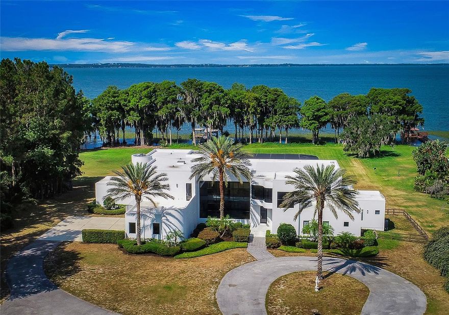 Welcome to this absolutely one-of-a-kind gated lakefront estate on Lake Harris and the Harris Chain of Lakes, a true architectural marvel that blends luxurious living with modern design across a sprawling 7,200 square foot custom-built home. Set on a gated 6.42-acre lot with no HOA, this property is a once-in-a-lifetime opportunity, offering complete privacy, breathtaking SUNSET views on the lake, and a private dock. From the moment you walk down the descending walkway into the dynamic great room, you're greeted by a 25-foot coffered ceiling, a cozy gas fireplace, and bold walls of glass that showcase the spectacular views. The main level is designed for luxury and convenience, featuring a large center island kitchen, a breakfast room, a family room with an expansive glass overlook to the waterfront, and a home theater equipped with a 9-foot custom Stewart screen and plus theater recliners. The luxurious master suite boasts a dramatic lighted ceiling, a circular glass block shower with a custom skylight, and a spacious walk-in closet. Upstairs, accessible by an ultra-wide center staircase or a custom steel spiral staircase, you'll find elevated walkways offering stunning views over the great room and out to the scenic grounds and Lake Harris. This level includes three additional bedrooms with walk-in closets, three full bathrooms, a 4-bed bunk room, and a glass-enclosed reading loft next to a built-in 24-foot library bookcase. The estate is an entertainer's dream with a screen-enclosed heated pool and spa, a summer kitchen, and a massive 60x80 pre-engineered red iron building perfect for storing collector cars, RVs, boats, or as a horse barn. This building is fully hurricane rated and features a 10-inch reinforced concrete floor strong enough for the heaviest equipment. With additional amenities like a three-car attached garage, a two-car detached garage, and two laundry rooms—one on each floor—this property doesn’t just promise a home but a new way of experiencing life by the water's edge. Whether you’re hosting grand scale events or seeking tranquility, this estate offers it all.