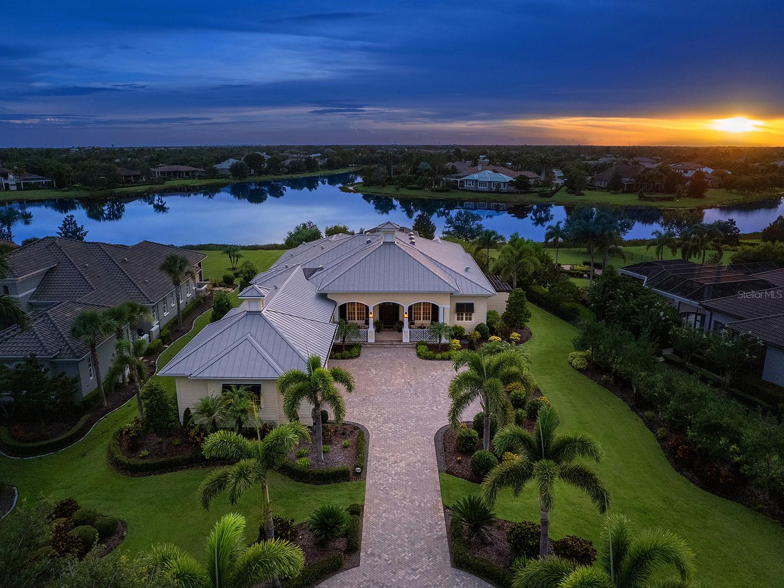 With stunning sunset views over the sparkling lake, this picture-perfect home in The Lake Club is truly a rare find in all of Lakewood Ranch! Built in 2016 is this completely custom home built by award-winning Nutter Custom Construction. Every detail in this residence has been meticulously selected for a luxurious yet functional lifestyle. Situated on 0.76 acres, this spacious lot affords you additional privacy, manicured landscaping, and over 80 feet of lakefront. In addition to the opulent upgrades inside, this home is outfitted with a rare METAL ROOF that offers up to 50 years of life, and IMPACT windows and doors throughout. From the spacious paver driveway surrounded by beautiful landscaping, you’re welcomed home to a charming front porch entry with Caribbean box railing and mahogany front door. The foyer invites you in with volume ceiling and custom painted wall to the open floor plan. The grand great room is truly the heart of the home and showcases vaulted ceilings with hand-stained wood beams, cozy electric fireplace, engineered hardwood flooring, custom built-in shelving, solar tubes for an abundance of natural light, and impact rated French doors to the lanai. Outdoors you’ll be immediately impressed with the western views of the lake, spacious gathering areas, custom ceilings, and colonnade screen enclosure. The heated pool and spa offers the ideal place to gather at one end around the gas firepit; or relax in the spa at the other end lakeside. Enjoy dinners alfresco from the outdoor kitchen expertly equipped with a Lynx grill, beverage refrigerator, and built-in ice chest cooler. Inside the chef will adore preparing meals in the gourmet kitchen complete with solid wood cabinetry including appliance bay and wine rack, quartz counters, central island with seating, and walk-in pantry with shelving. The high-end appliance suite includes a Jenn-Air paneled refrigerator, Wolf built-in microwave and wall oven, KitchenAid warming drawer, Miele dishwasher, and Bosch induction cooktop with flex bridge burners that make cooking a breeze. Dine in the formal dining room with designer chandelier, or in the casual dinette with built-in cabinetry. When you’re ready to end the day in relaxation, retreat to the primary suite with vaulted ceiling, dual custom walk-in closets, separate seating area, and private lanai with remote-controlled shades. Your primary bath is a sanctuary with dual split vanities, limestone tile, walk-in shower with bench, soaking tub, and private courtyard. Guests will enjoy their own luxe accommodations in the second primary bedroom with private en-suite bathroom and walk-in closet that is ideal for long-term guests or could be used as a bonus room; or the two additional guest bedrooms with walk-in closets and jack & jill bathroom. Rounding out this one-of-a-kind residence is a study with custom built-in shelving and desk, laundry room with plentiful storage, mudroom entry with 3 closets, and guest powder bathroom. Don’t forget about the oversized 3-car garage with fans to keep cool, custom storage cabinetry and workbench, and air-conditioned Florida basement perfect for storage. The Lake Club is an exclusive premier, gated community with a Grande Clubhouse, restaurant, pool, tennis, pickleball and more.