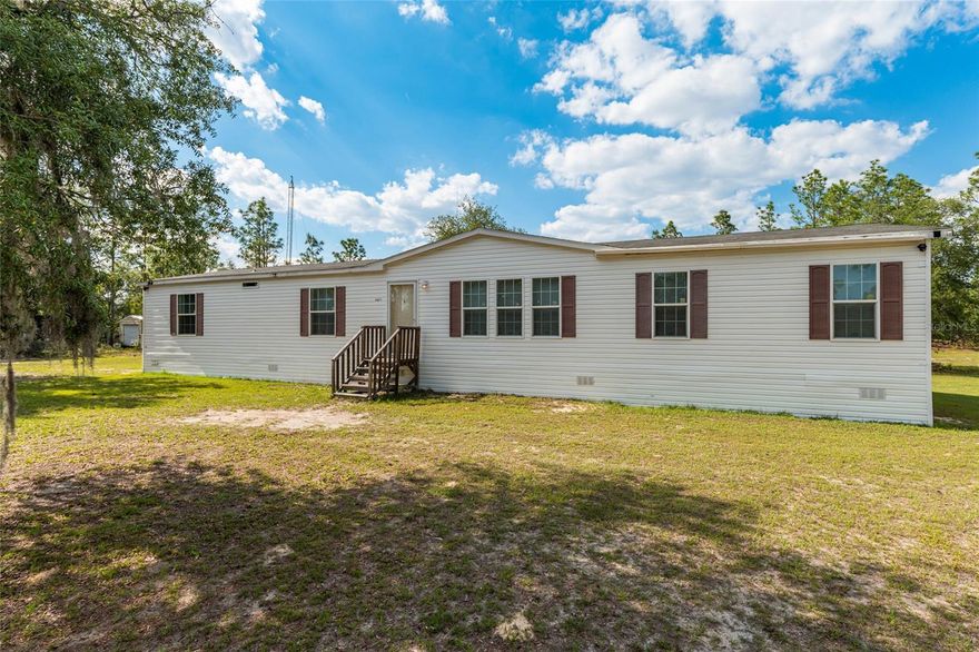 Step inside this double-wide manufactured home and be immediately impressed — tray ceilings, crown molding, and a sweeping view of your own land right through the back door make it feel anything but ordinary. The open feel continues into the large kitchen, complete with a center island, decorative range hood, and a cleverly concealed pantry designed to blend seamlessly with the cabinetry.  With 4 bedrooms and 3 bathrooms, there is room for everyone. The primary suite offers a private retreat, while a Jack-and-Jill configuration serves two of the additional bedrooms — all featuring built-in closets. A fourth bedroom provides flexibility for guests, a home office, or whatever suits your needs.
There is a short drive, less than half a mile, on a regularly scheduled, county-maintained limerock road to reach the property, which sits on 2.5 acres across two taxable lots, with the ability to split and add future value. Outside, the vinyl siding has been extended to ground level for a clean, polished look with no visible skirting. Generator wiring is already in place, making it easy to add a whole-home generator at your convenience. Gutters are currently installed on one area
, with all remaining materials on hand to complete the full installation.   A rare find with space, style, flexibility, and peace — don’t miss it!
