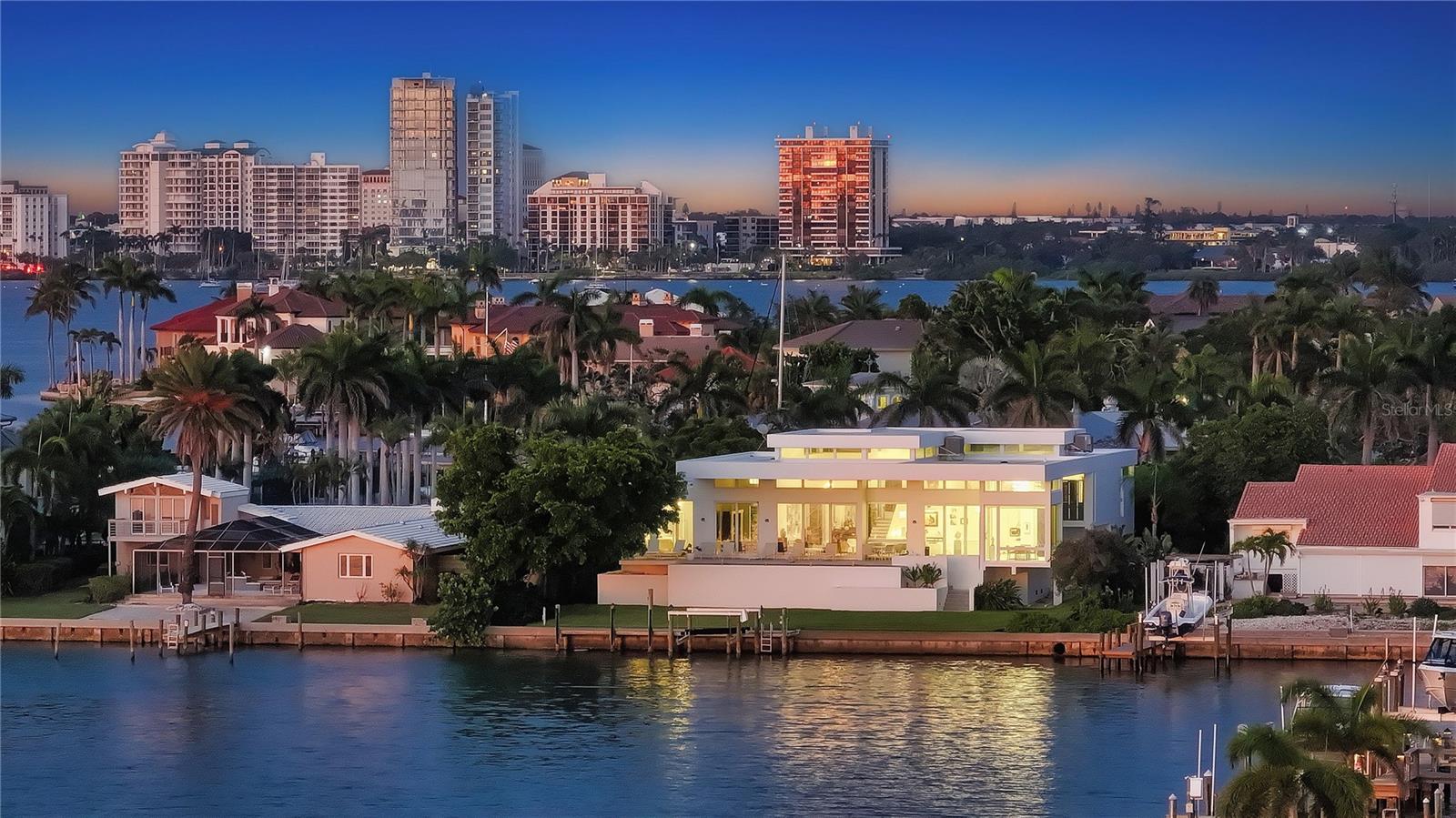 Located on the pristine shores of Sarasota Bay, this striking contemporary Bird Key residence offers a seamless fusion of bold architectural design and elevated coastal living. Designed by renowned architect Jonathan Parks and completed in 2014, the home spans over 4,100 square feet across two living levels. Every inch reflects a refined aesthetic, with soaring ceilings, luminous marble floors, and expansive walls of glass that frame breathtaking water views and spectacular sunsets. A circular driveway introduces the home, guiding you through manicured landscaping to a dramatic marble and glass-framed foyer. Once inside, the open-concept layout unfolds in impressive fashion. The expansive living/dining room is anchored by 20-foot ceilings and clerestory windows that flood the space with natural light, while sliding glass doors blur the line between indoors and out. Perfectly suited for entertaining or everyday living, the main living area flows effortlessly into a show-stopping kitchen equipped with sleek custom cabinetry, a waterfall-edge island, gas Wolf range, walk-in pantry, built-in planning desk, and wet bar. The main-level primary suite is a tranquil retreat, featuring dual walk-in closets, separate en-suite bathrooms for him and her, and a private study offering a quiet place to relax. Upstairs, a spacious loft functions beautifully as a game room, media lounge, or additional workspace, while two well-appointed guest suites offer walk-in closets and private balcony access overlooking the surrounding treetops and waterway. Outdoors, the setting is equally remarkable. With 100 feet of direct bay frontage, the backyard invites endless possibilities for recreation. A long lawn is bordered by a flowing fountain feature, offering the perfect canvas for creating your own resort-style pool and al fresco entertaining space. Boating enthusiasts will appreciate the newer dock equipped with a 10,000-pound lift, allowing for easy, unobstructed access to Sarasota Bay and the Gulf of Mexico beyond. Situated on the exclusive barrier island of Bird Key, this modern residence places you just minutes from Sarasota’s vibrant downtown, the fine dining and upscale boutiques of St. Armands Circle, and the immaculate Gulf sands of Lido, Longboat Key, and Siesta Key beaches. Cultural venues like the Ringling Museum and Sarasota Opera are nearby, along with premier yacht clubs and marinas. Offering a rare blend of modern design, waterfront serenity, and proximity to the region’s finest amenities, this home captures the very essence of an oceanside luxury lifestyle.