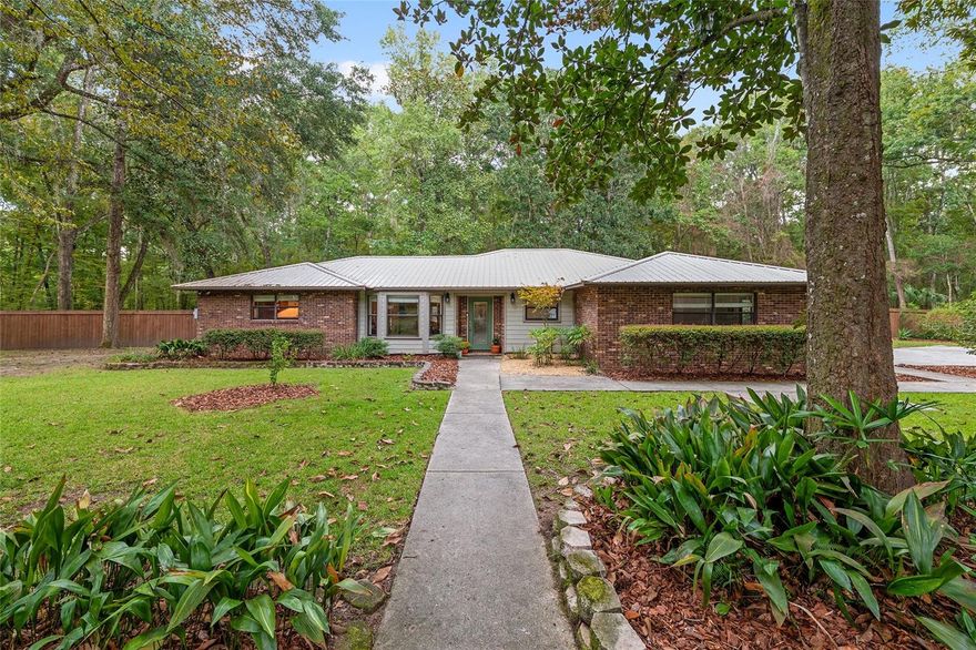 Discover the perfect blend of tranquility and convenience in this beautiful home nestled on nearly an acre in Marmaduke West off scenic Millhopper Road. Surrounded by nature, this property offers unmatched privacy, a serene backdrop of mature trees and natural beauty.  Approaching the property and the cul-d-sac, you will immediately notice the lovely front landscaping, extra-long driveway, exterior brick accents, and newer metal roof on this 4-bedroom, 3 bath home.  Inside, you’ll find light-filled living spaces designed for comfort and a connection with the outdoors.  Entering the foyer, you are greeted by gleaming wood floors which flow throughout the entire home, no carpeting.  The large great room features a stacked travertine stone wall encompassing shelving, built-in TV, and a unique ethanol Eco Smart fireplace, a clean burning alternative to wood burning or gas.   To the right of the foyer is a spacious office area, large enough for several desks.  The open floor plan highlights an amazing contemporary kitchen featuring stunning floor to ceiling wood cabinets, quartz counters, SS appliances, and unique designer tile backsplash that sets off the entire area.  A cozy breakfast area completes the kitchen and a more formal dining area bridges the kitchen with the great room.  The split bedroom plan features a large owner’s suite with two sets of french doors to the patio area and 2 amazing barn doors leading to the walk-in closet and the owner’s bath.  The bath is newly renovated and resembles a luxury spa with double wood vanity with quartz counter and a striking glass enclosed shower with floor to ceiling stone like tile.  At the opposite side of the house are two large guest rooms and hall tub bath with two pedestal sinks.  A unique feature of this special home is a 420 sq. ft. in-law suite with full kitchen, plenty of space for a seating area, and comes complete with a murphy bed.  An adjacent full bath features a jetted tub. The in-law suite can be accessed either thru the home or thru a private entrance. Enjoy your morning coffee or unwind in the evening on the expansive screen porch which overlooks an over-sized open patio. Majestic trees grace the private fully fenced yard with the added bonus of two storage sheds, plenty of room for all your gardening tools and extras.  Whether you’re seeking a peaceful retreat or a convenient home base close to University of Florida, shopping, and restaurants, this property and Marmaduke West, with no HOA, delivers both.  Experience the best of both worlds—quiet, scenic living with easy access to everything you need.  Move-in ready with a 2022 metal roof and 2025 HVAC.