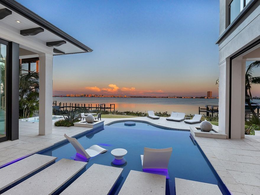 Set against Sarasota’s coveted bayfront in Lido Shores, this extraordinary waterfront estate captures sweeping views of Sarasota Bay, the downtown skyline, Ringling Bridge, and City Island, offering an unparalleled luxury lifestyle with effortless access to boating, beaches, and downtown Sarasota. More than a residence, this is a statement property—an iconic example of West Indies–inspired architecture executed with exceptional craftsmanship and modern sophistication. A private gated entry introduces a home where over $1 million in enhancements and more than 2,000 hours of custom design are evident throughout. Advanced smart home automation allows seamless global control, blending innovation with refined living. The custom front door opens to a dramatic foyer and a great room defined by a coffered ceiling, media wall, linear fireplace, and expansive bay views. The dining space features a quartz-topped bar with wine storage and flows into a chef’s kitchen appointed with Thermador appliances, a large floating island with quartz waterfall edges, and a walk-in pantry with an additional Bosch refrigerator. The primary suite is a tranquil sanctuary with floor-to-ceiling glass, sliders, and breathtaking water views. Two custom walk-in closets complement a spa-inspired bath featuring floating vanities, a soaking tub, a custom multi-head shower, and sliders leading to a private Zen garden with an outdoor shower. A sculptural staircase and glass-enclosed wine wall lead to an upper-level loft with a wet bar and waterfront terrace. A refined office with custom built-ins and four en-suite guest bedrooms, each with custom closets, projector screens, and private balconies, ensure comfort and privacy throughout. Outdoors, the estate transforms into a resort-style waterfront retreat. An infinity-edge pool with sun shelf, in-pool table, and cascading spa anchors the setting, surrounded by multiple seating areas, a firepit, and an outdoor shower. The fully equipped outdoor kitchen—featuring a smoker, grill, wok, and side burner—makes alfresco entertaining effortless, while a spiral staircase leads to an upper terrace with panoramic views. The newer deepwater dock is designed for serious boating, offering a 20,000-pound boat lift, jet ski lift, water and power, ambient lighting, and a dedicated fishing area. With direct access to Sarasota Bay just steps from the backyard, this property delivers exceptional boating and waterfront living. Additional features include a three-car garage, two laundry rooms, an elevator, a garage drop zone, and a hidden utility closet. The home is fully integrated with Control4 automation, security cameras, a 48-kilowatt generator, 20-kilowatt battery backup, high-end audio-visual systems, automated shades, circadian lighting, outdoor theater, app-controlled pool and irrigation, Wi-Fi-enabled appliances, EV charger prewiring, Starlink backup internet, motion-activated lighting, and voice control via Josh AI. Lido Shores is one of Sarasota’s most sought-after and architecturally significant waterfront communities, offering residents exclusive access to the north end of Lido Beach and close proximity to St. Armands Circle, Longboat Key, City Island, and downtown Sarasota. This remarkable bayfront estate is an architectural triumph, designed for sophisticated entertaining, advanced living, and extraordinary waterfront luxury.