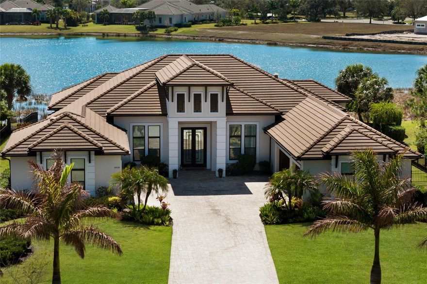 A stately arrival sets the tone for this exceptional custom residence, thoughtfully positioned on over one acre within the gates of The Concession. Blending modern luxury with architectural refinement, the home offers a rare combination of privacy, elegance, and timeless design in one of Lakewood Ranch’s most exclusive communities. Custom double entry doors welcome you into a light-filled interior where engineered hardwood floors anchor the main living spaces and a dramatic wall of sliding glass doors frame serene water views, seamlessly connecting indoor and outdoor living. At the heart of the residence, the chef’s kitchen is both polished and highly functional, appointed with Monogram and Café appliances and anchored by a generous statement quartz island that serves as both focal point and gathering space. Designed to flow effortlessly into the great room and outdoor living areas, the layout supports both elevated entertaining and comfortable everyday living. Architectural elements define the great room, where a custom-designed accent wall showcases intricate detailing, a sleek linear fireplace, and an oversized mounted television, creating a striking focal point that balances warmth with modern design. Tray ceilings throughout further enhance the home’s architectural character, while curated designer lighting, plantation shutters, and a newly installed outdoor surround sound system underscore the thoughtfully executed craftsmanship. The primary suite offers a private retreat of generous proportions, complete with a sitting area and dual custom walk-in closets. The spa-inspired bath features expansive dual vanities, a soaking tub, and a dramatic walk-through shower with triple shower heads, including an overhead rain feature delivering a resort-caliber experience. Two additional guest suites each offer beautifully appointed en-suite baths with elevated design selections tailored for today’s discerning buyer. A private office with oversized windows invites abundant natural light, while the formal dining room is enhanced by a dramatic wine display that serves as a striking focal point for elegant entertaining. A spacious bonus room provides flexible space for recreation, media, or additional guest accommodations. Outdoors, the experience continues with a large pool and spa, expansive covered lanai, and a nicely appointed outdoor kitchen, creating a true resort-style setting. The five-car garage provides exceptional capacity for the automotive enthusiast and additional storage needs, while a fenced yard allows for secure outdoor enjoyment. Constructed with hurricane impact doors and windows throughout and supported by a whole-house generator, the home offers peace of mind with uncompromising quality. This residence masterfully blends functionality with a refined, designer-forward aesthetic, delivering privacy, security, and elevated living. Located just minutes from UTC, Waterside dining and shopping, and the Gulf Coast’s renowned beaches, it offers proximity to everything while maintaining an atmosphere of serene exclusivity. A distinguished offering for those seeking elevated luxury and the true Concession lifestyle.