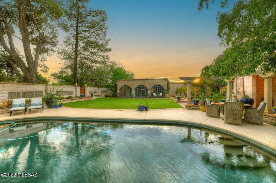 Like so many beautiful homes, the story begins as you enter hand forged iron gates. A gracious compound with a classic adobe home, a guest casita with wall of french doors,  columned terrace, pool, and all  nestled around green grass, magnificent trees, and splashes of flowers. It is a magical place that causes everyone to linger.  ''Zaguan'' which means  entrance in spanish, is a charming way to enter this special home.   A seguay through glass doors to the north patio where unobstructed mountain views prevail.  The great room can only be called breathtaking.  Bringing the views in at every turn it is large, light,  airy,and yet  so inviting.   The formal dining room is just as it should be..classy, beautiful lighting, and designed for long conversations around the dinner  table. A gourmet kitchen with a fireplace/pizza oven, concrete counters, and anchored by a magnificent wolf stove. It is done right. 
One of my favorite rooms is a library that was designed with every  attention to detail....picture custom cabinetry, room for a magnificent office, and still a reading area cozied up to the fireplace.   A split bedroom plan,the master shares the same magnificent windows and views as the rest of the home.  A gracious fireplace and seating area completes the suite. 
The guest casita with its brick floors and full bath is so welcoming.   I could go on and on.  As a Tucson native, I love finding a home that defines my city.