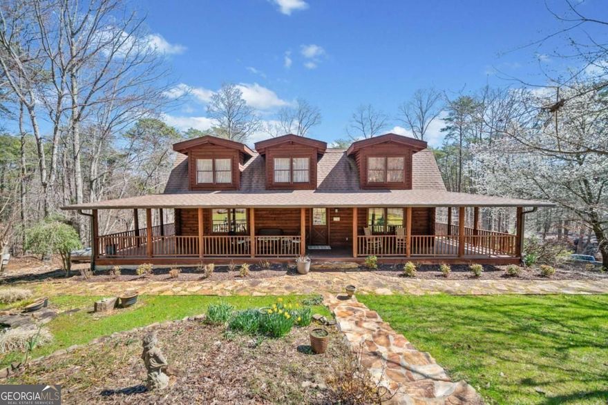 Cape Cod-style log home tucked away on 12.54 acres. It is built with high-quality materials and features rich heart pine wood floors, vaulted beamed ceilings, and a welcoming wraparound porch on all four sides. The kitchen has granite countertops, a white farmhouse sink, a spacious island, and a cozy second fireplace. The two-story great room features a brick fireplace, vaulted ceilings and exposed beams. The main-level primary suite provides convenience with an attached bath, which has been recently updated to include a handicap-accessible shower. The upper level has three additional bedrooms, each of which shares a hallway bathroom. Downstairs, the unfinished basement includes of a full bath with the potential for extra living space. Outside, a separate screened-in party pavilion is perfect for gatherings, while the barn workshop, complete with second-floor storage and an additional equipment outbuilding, provides ample space for hobbies and storage. A professionally installed whole-house generator ensures peace of mind. Lush landscaping, blooming perennials, and a small creek running through the property add to the charm of this one-of-a-kind escape.
