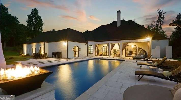 Welcome to a rare offering in luxury real estate-an extraordinary custom-designed residence nestled behind double-entry gates on 2.68 acres of pristine privacy. Located just moments from Lake Lanier, this fully furnished estate redefines refined living with unmatched style, function, and resort-inspired amenities. From the moment you enter the grand foyer, soaring ceilings, walls of glass, and architectural artistry capture your attention. A dramatic formal dining room and richly paneled cigar/library lounge set an elegant tone, while a two-sided fireplace anchors the expansive living room and flows effortlessly into the gourmet chef's kitchen-outfitted with an ILVE Majestic dual-fuel range, quartzite countertops, a sun-soaked breakfast nook, and sprawling island designed for entertaining. The main-level primary suite is a personal retreat featuring a spa-like bath with a steam room, essential oil soaking tub, and dual custom closets. Upstairs, four spacious en-suites impress with bespoke finishes and a gallery-like overlook framed by imported cast iron balusters. A private guest house, connected by a covered porte-cochere, includes a full luxury kitchen, walk-in pantry, and laundry-perfect for long-term guests or multi-generational living. Outdoors, enjoy the spectacular heated pool and spa, complete with waterfall features and fire elements, surrounded by lush landscaping and proximity to a private boat slip (just 0.35 miles away) completes the dream. Also includes a theater room and pet grooming station (not pictured). Offered fully furnished and move-in ready.