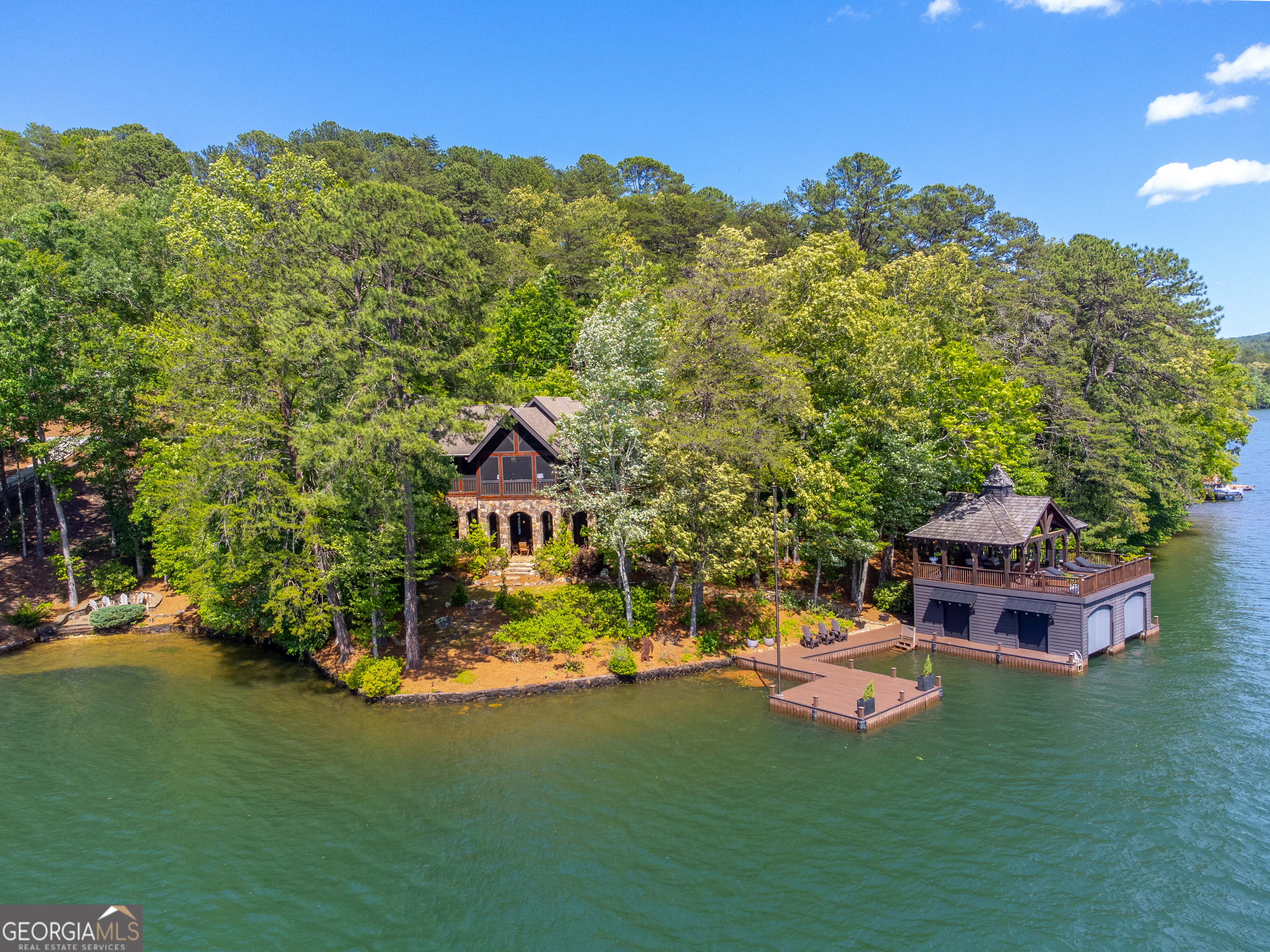 This spectacular lakeside estate rests on the shores of one of North Georgia's most exclusive deep water mountain lakes. Perched on a gently sloped point lot with 300' of shoreline and expansive westerly views, this classic mountain home on Lake Burton offers 4 bedrooms, 4.5 baths, plus a bunk room. The great room features a masonry stone fireplace, heart pine flooring, vaulted ceilings with Douglas fir timber frame construction, and French doors that reveal sweeping lake and mountain views through the screened porch. An open floor plan connects the kitchen-equipped with a Thermador 5-burner gas cooktop, KitchenAid wall oven, warming drawer, ice maker, 36" fridge and refrigerator drawers-to the main living area and dining area. The lakeside screened porch showcases mountain sunset views while providing easy flow for entertaining with a generous lounge area and adjacent dining area with built-in grill and fireplace. The private main-level owner's suite provides lake views, a lakeside deck, and a well-appointed bath with separate vanities, travertine tiled shower, jetted tub, and walk-in closet. A guest suite with lake views and en-suite bath, powder room, laundry room, and two-car garage complete the main floor. The terrace level offers a lodge-style family room with 10'+ ceilings, a stone fireplace, billiards area, and a custom bar featuring a heart pine countertop, sink, under-counter fridge, ice maker, dishwasher, and built-in microwave. Two guest bedrooms, each with lake views and private baths, flank the terrace level living space-one with an adjoining bunk room, the other with access to a covered lakeside patio. Outside, a covered stone patio includes a dining area, built-in smoker, and power burner with a pathway leading to a lakeside firepit in one direction and a two-stall, two-story boathouse and swim dock in the other. The boathouse sits on deep water and enjoys panoramic views of Billy Goat Island and a front row seat to the Lake Burton 4th of July fireworks. Two jet ski lifts are mounted on the side of the boat house and metal security doors protect boats from the weather year round. This special point lot is gently sloped with minimal steps from the terrace level to the boathouse and enjoys the advantage of fully paved access by road. This home was built by Chris Clay in 2006 and has been meticulously maintained.