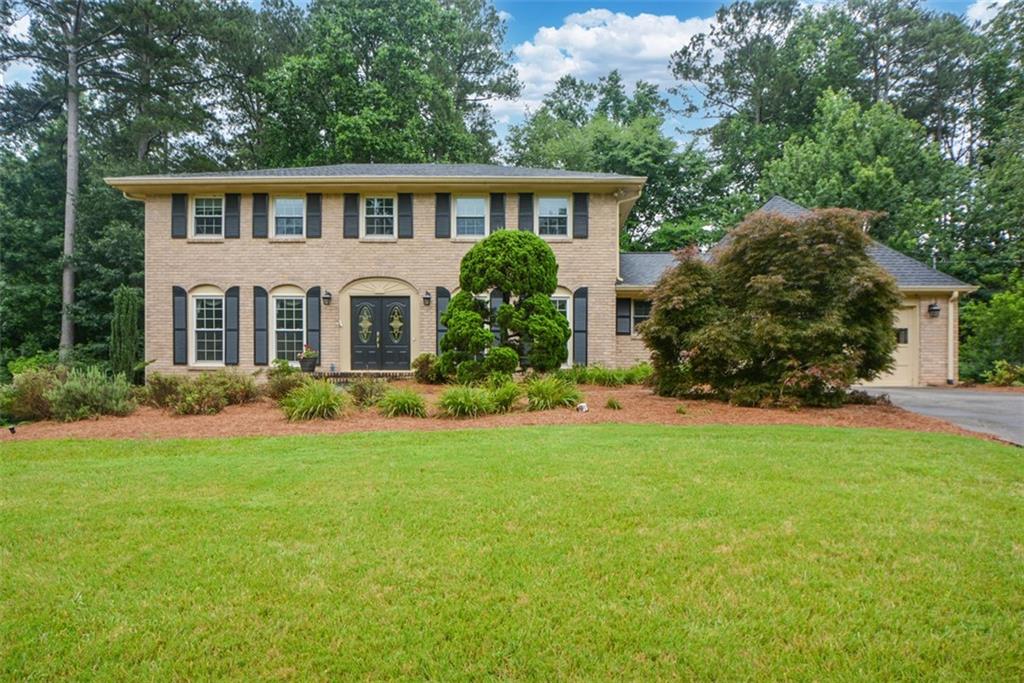 Brick facade residence featuring a two-story design with a double-door entry, black window shutters, an attached two-car garage, and a lush lawn with mature landscaping