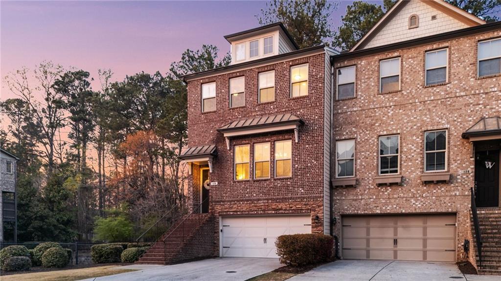 End unit brick townhouse featuring a two-car garage, dormer window, and covered entry with open space next to it and woods behind.