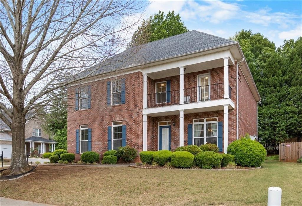 Brick facade residence featuring a two-story columned front porch with a balcony, blue louvered shutters, and established perimeter shrubbery