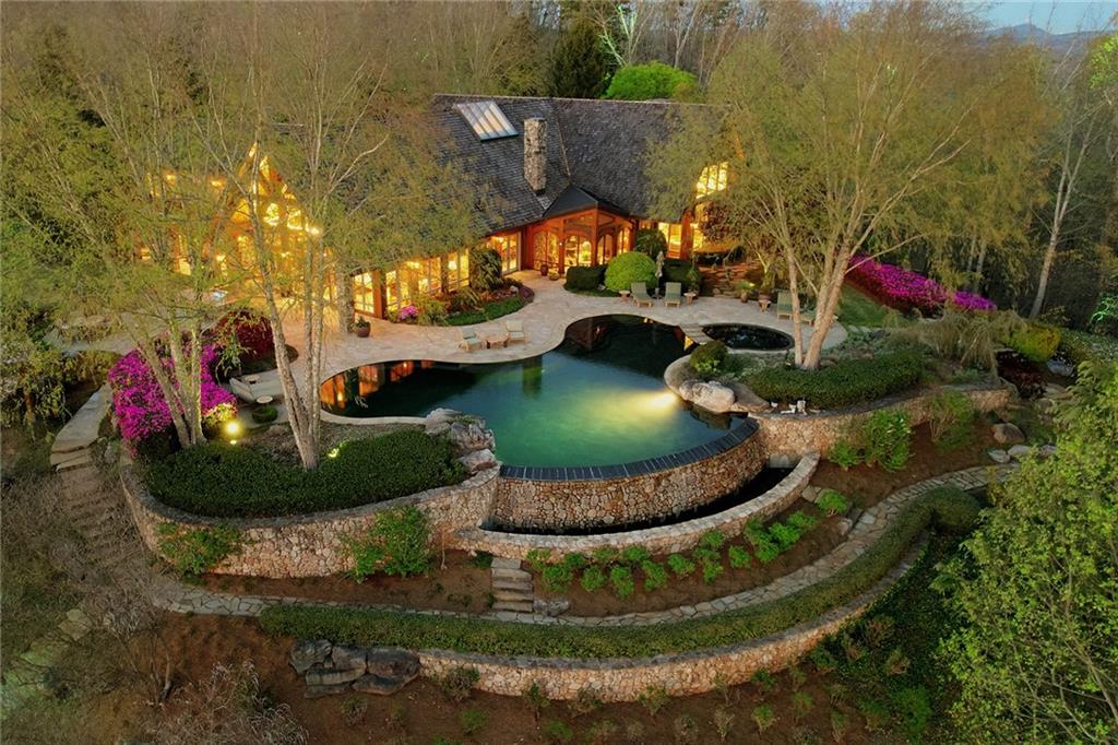 Welcome to this one-of-a-kind, custom-built retreat in North Georgia, surrounded by a lush landscape and spectacular mountain views! Exquisitely designed by William Stewart of Stewart Mohr Designs, this private estate is sure to take your breath away from the moment you enter through the winding, gated drive. The great room exudes warmth, volume and strength, featuring a wall of windows, a leather entryway wall, wide-plank black walnut flooring, a soaring 24’ vaulted ceiling with on-site hand-cut Douglas fir trusses and one of six dry-stacked or river rock floor-to-ceiling fireplaces. A custom motorized TV life console hides the TV when not in use
and provides a graceful separation between the living and dining areas. The fireside gourmet eat-in kitchen features a stone fireplace, custom dark walnut cabinets, a massive kitchen island with seating, Viking and Sub-Zero appliances, a pot filler, a prep sink, exotic leathered granite countertops and radiant heat stone flooring. A huge walk-in hidden pantry with an appliance garage features a full freezer for extra storage. Natural light pours into this space from the vaulted ceiling with skylights. An adjacent, inviting game room off the kitchen impresses with picture windows and a full bar looking out to the outdoor kitchen and spacious stone patio. The primary suite – or, more aptly, the owner’s wing – of the home offers a generous fireside master bedroom
with a morning bar and a fireside sitting room with built-ins. Relax in the spa-inspired bathroom outfitted with a walk-in shower and a built-in bathtub featuring captivating views of the surrounding landscape. This area is created for comfort with radiant heat flooring, dual separate bathrooms, a custom walk-in closet, a separate walk-n cedar closet, a private balcony and a private owner’s office. Built for entertaining, this residence boasts a guest wing on the main level, complete with two powder rooms, a handsome den and a home manager’s office – both featuring reclaimed pine walls and cabinets, a spacious gym,
an enormous mudroom and a laundry room with a full bathroom and extensive storage. Through the spacious hall highlighted by a wall of windows that floods the space with natural light, discover a two- story floating black walnut staircase leading to the second level. Designed as a statement piece, the stairs are supported by an iron sculpture and lead to three large junior suites. Each bedroom includes generous closets and private bathrooms, and open onto the fabulous second-level balcony. The exquisite craftsmanship of the home itself is nothing compared to the true star of the estate – the outside living space. This isn’t just any backyard! This paradise serves a second living area, with a well-outfitted outdoor kitchen, multiple patios and an outdoor living room featuring a stone fireplace. The resort-style infinity saltwater pool includes a Pebble Tec finish and must be see in person to appreciate. In addition to beautifully landscaped grounds, the property includes a fabulous potting room attached to the three car garage with abundant storage, walls of windows and a half bathroom, plus a charming greenhouse, gardens galore and raised planting beds. The three-car oversized garage is temperature-controlled with slate floors and provides the perfect place for all your automotive needs. Absolutely no expense or detail has been overlooked! A private paradise lovingly
crafted down to the last detail, this home’s landscaped walkways, gardens and enchanting lookouts are designed to captivate with color and interest all year long!  It simply would not be possible to build a property of this caliber with these grounds anywhere close to today's pricing.